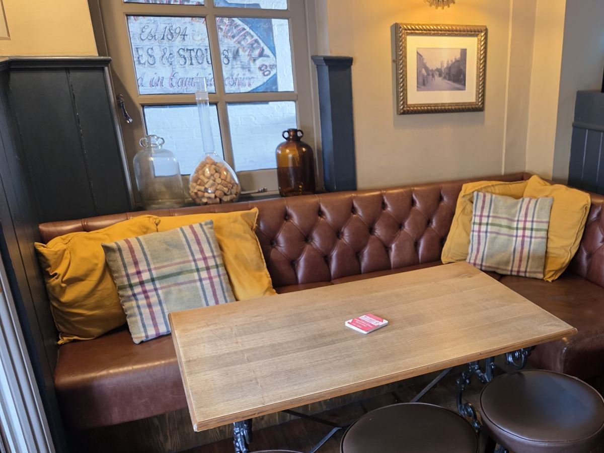 A tufted leather banquette with mustard and tartan cushions beneath a window painted with old brewery lettering