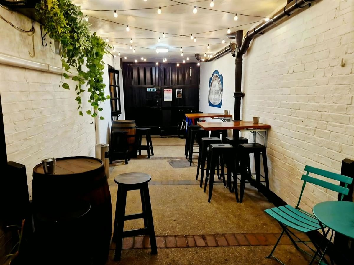 The passage to the courtyard at night, lit by festoon lights, with barrel tables, high stools and trailing plants