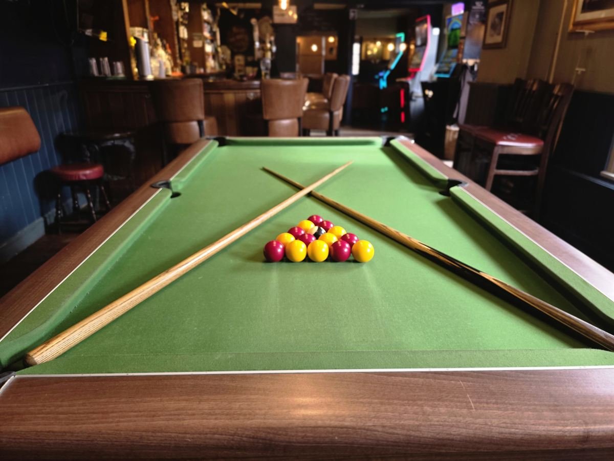 Two cues crossed over a racked set of red and yellow balls on the pool table, with the bar behind